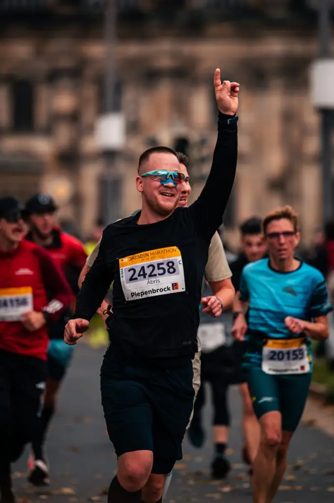 Jubelnder Läufer mit Startnummer 24258 beim Dresden Marathon 2025 vor der Semperoper, mit erhobener Hand und blauer Sportbrille