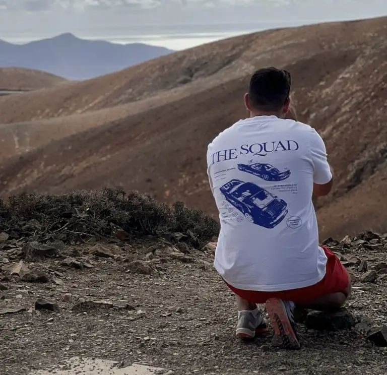 Patrick Baumgart, Gründer von Kicap Studio, kniend von hinten auf einem Aussichtspunkt in einer kargen Vulkanlandschaft mit Bergen und Meer im Hintergrund, weißes JP-Performance T-Shirt und rote Shorts.