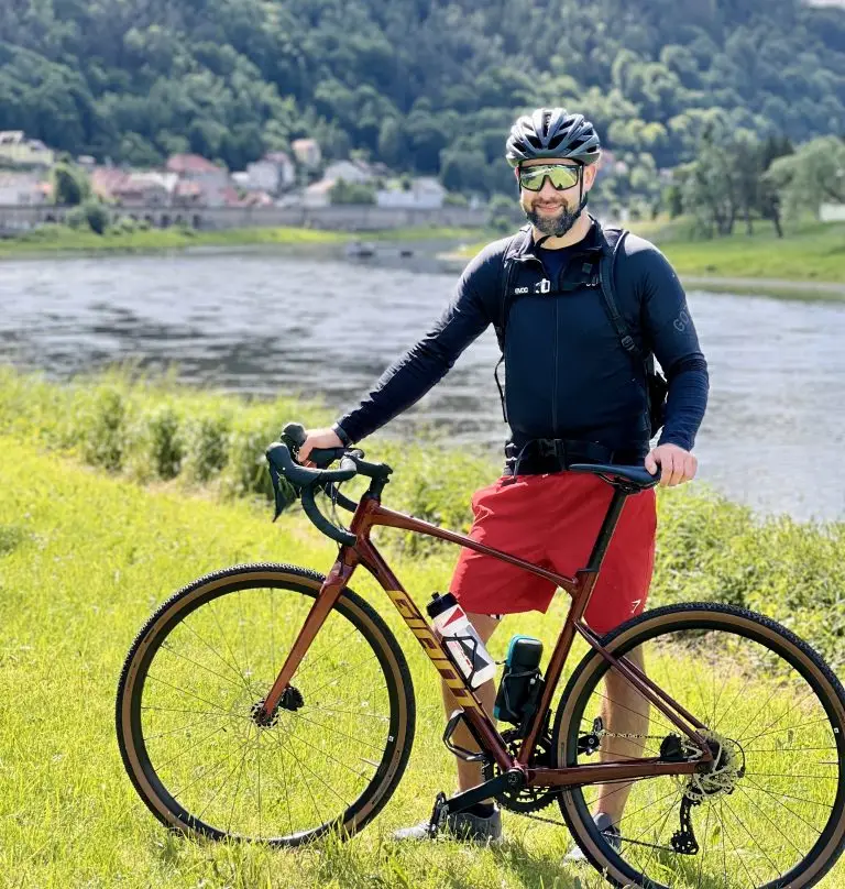 Patrick Baumgart, Sport- und Autofotograf von Kicap Studio aus Dresden, mit Gravelbike, Fahrradhelm und Sportbrille an einem Flussufer mit grüner Landschaft und Ortschaft im Hintergrund.