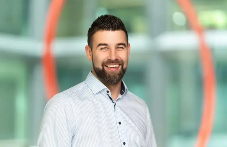 Patrick Baumgart, Sport- und Autofotograf und Gründer von Kicap Studio aus Dresden, lächelnd im Business-Portrait mit hellblauem Hemd vor modernem, farbigem Hintergrund.