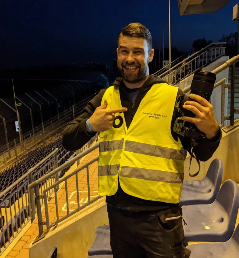 Patrick Baumgart, Sport- und Autofotograf von Kicap Studio aus Dresden, mit gelber Porsche Sports Cup Presseweste und Kamera in der Hand auf einer Tribüne am Lausitzring am Abend.